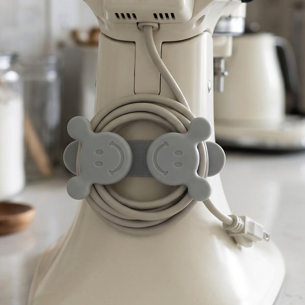A gray silicone cord organizer with two smiling faces, holding a coiled light gray appliance cord on a white stand mixer.
